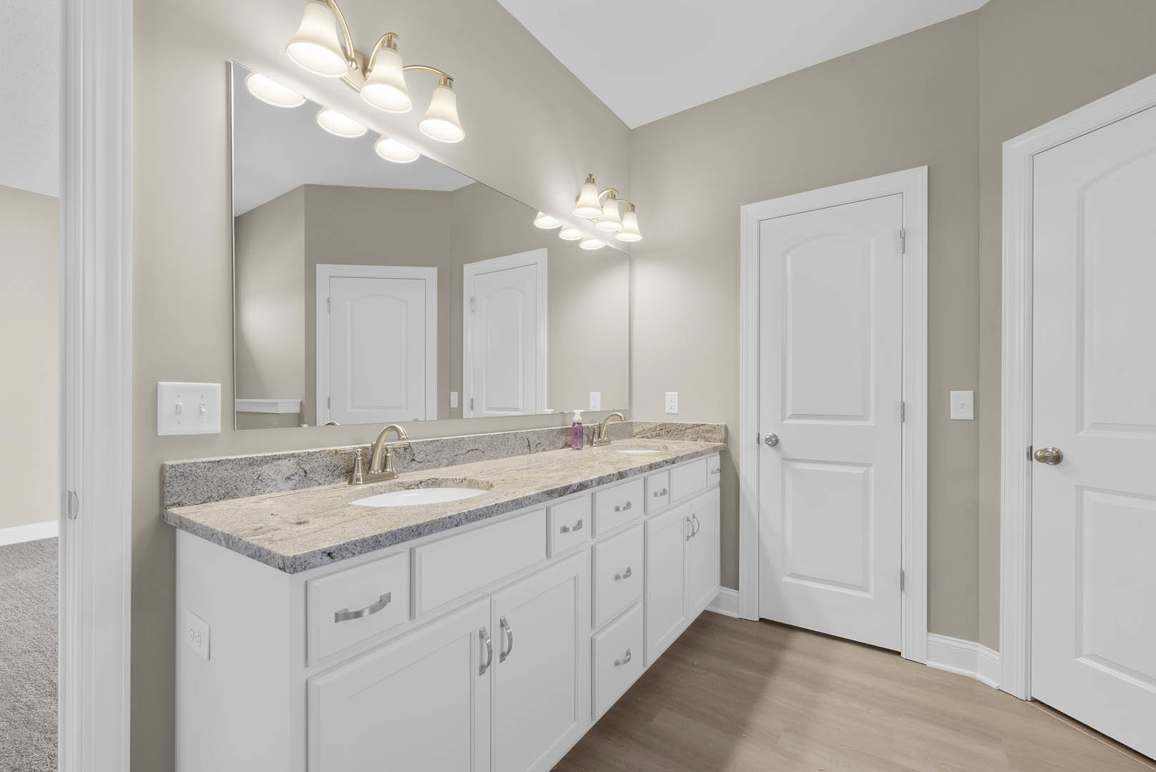 Bathroom with double white sinks, wide frameless mirror, white cabinetry, silver hardware, and a white door with silver handle.