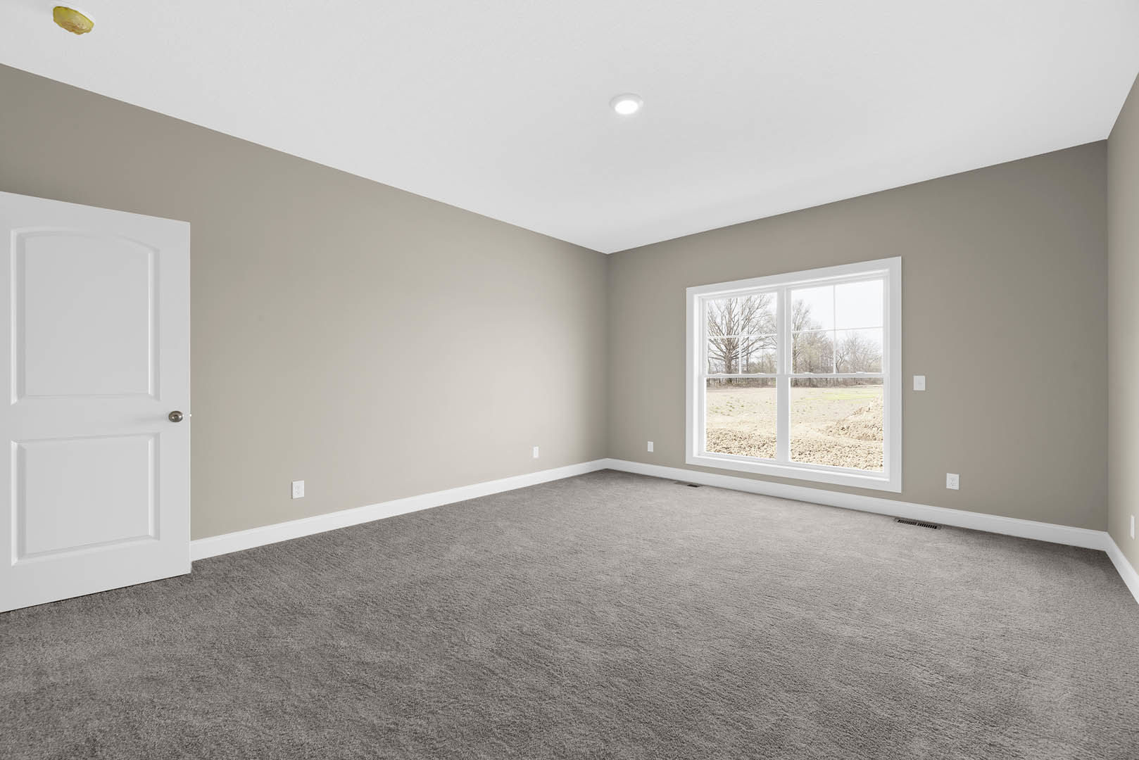 Bright room with grey carpet, white walls, white ceiling with recessed light, large window overlooking field and trees, white door with molding