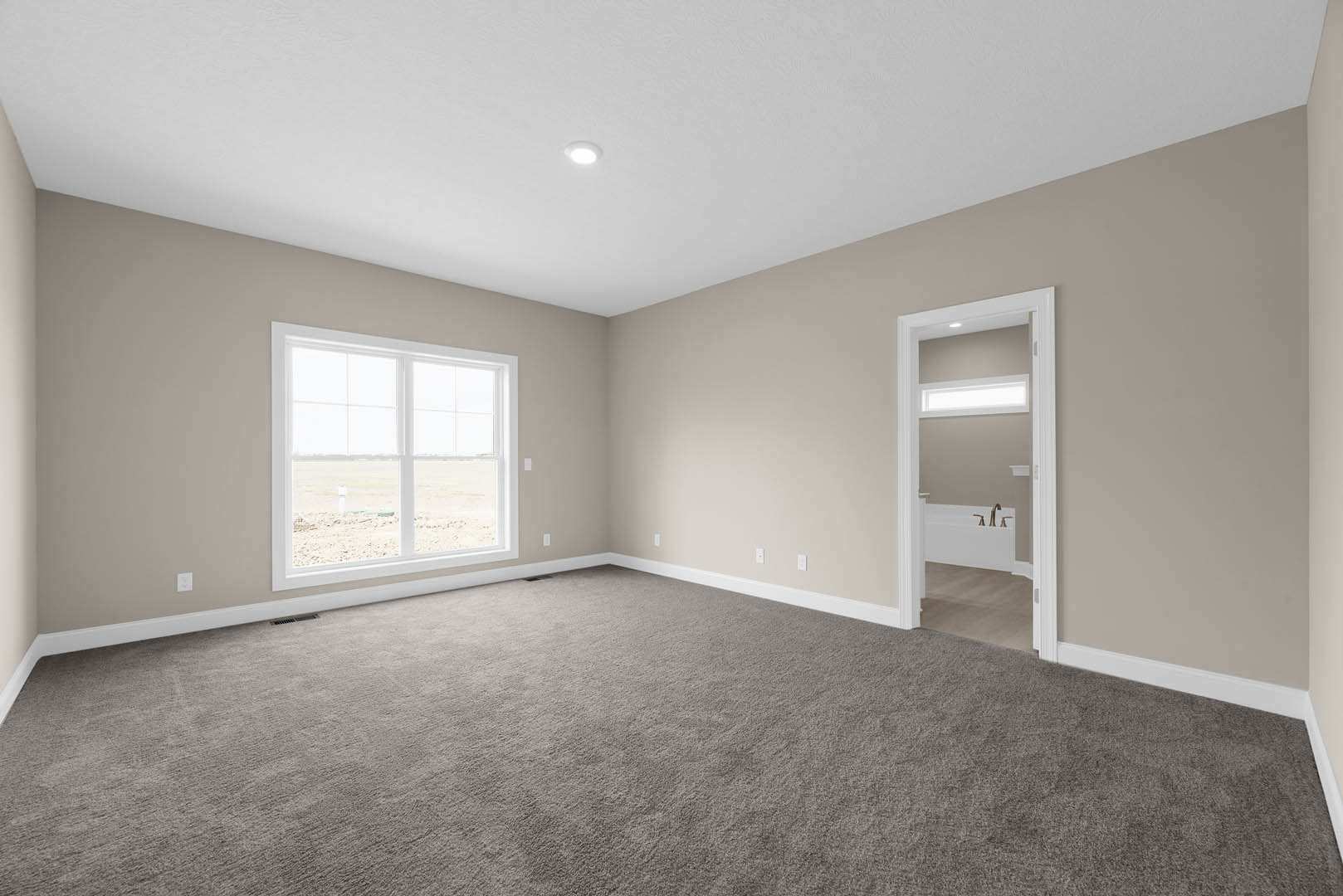 Bedroom with beige carpet, open white door, large window overlooking grassy field, white ceiling with recessed light fixture, partial view of bathroom featuring freestanding tub