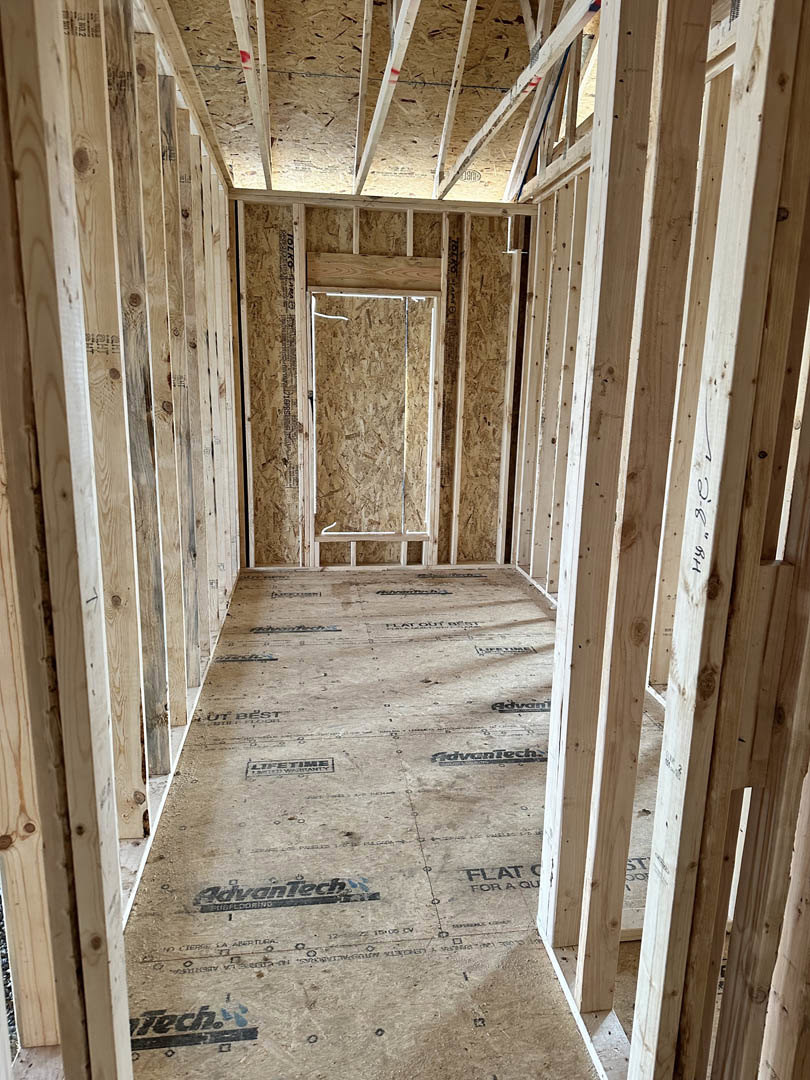 Room under construction with exposed wood framing, long hallway featuring wood beams, wood paneled wall with white trim, close-up of construction logo, partial view of roof