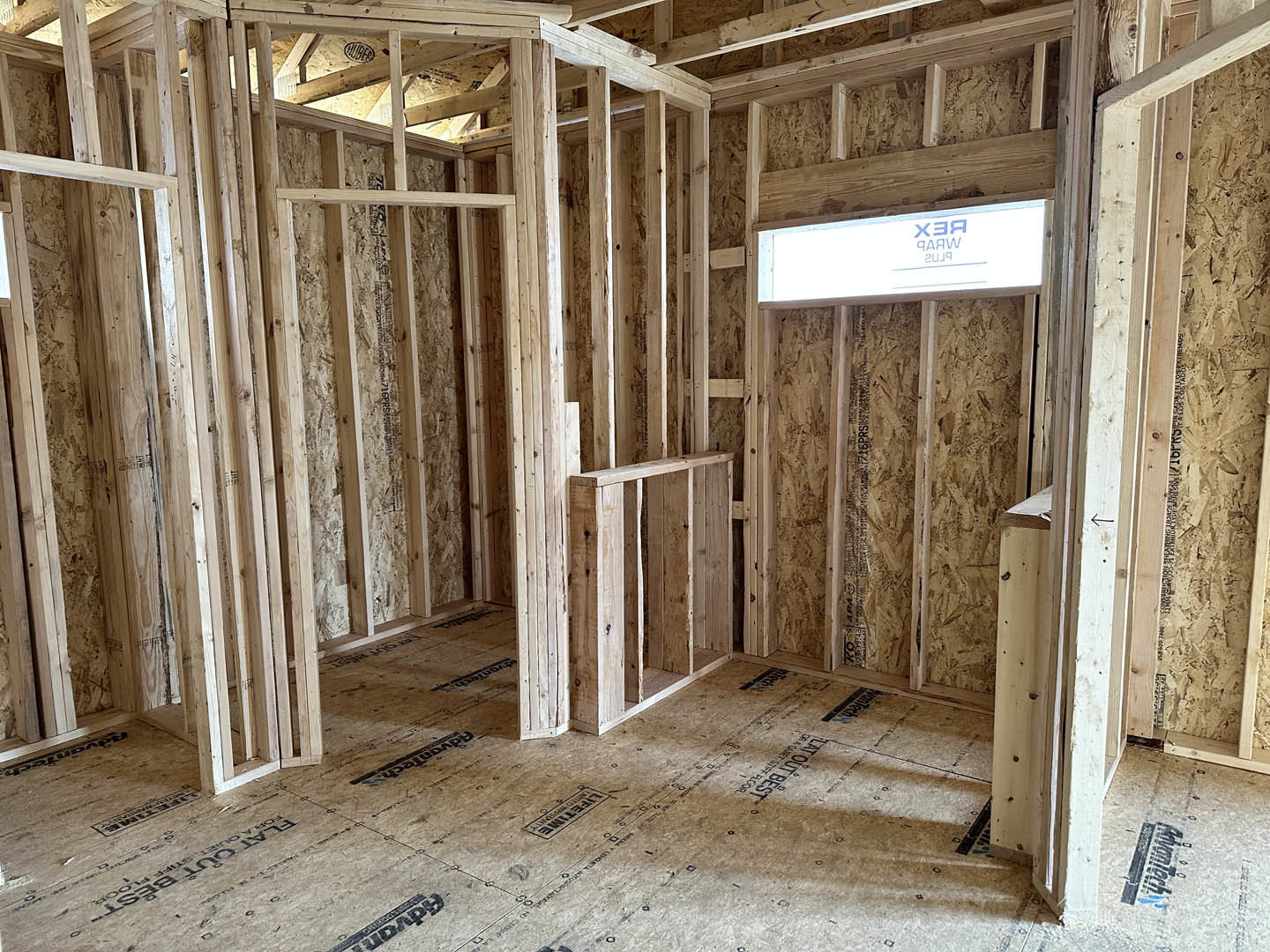 Room under construction with exposed wood framing, beams, and unfinished walls, blue sign visible in background, wooden shelf and plank flooring.