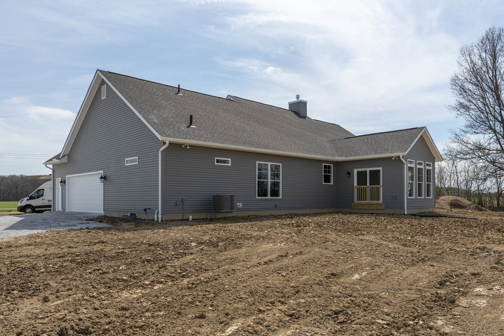 Modern house with light siding, white-framed windows, and a dirt field in the foreground; white van parked beside the home, trees and cloudy sky in the background