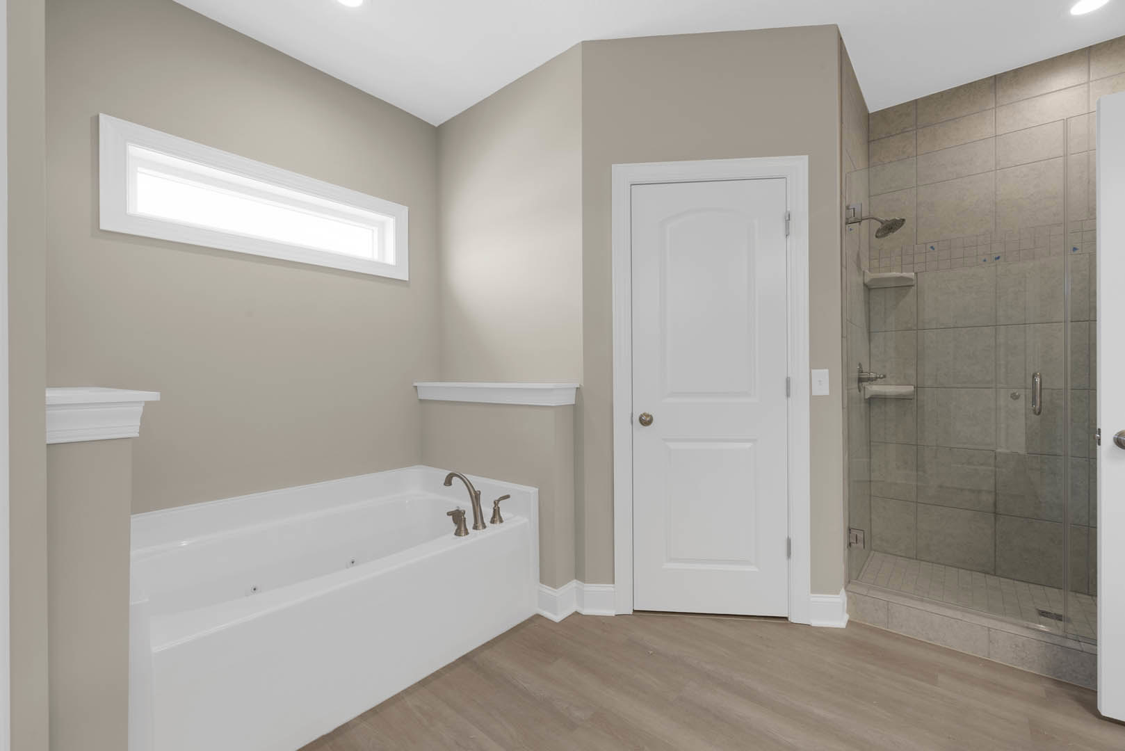 Bathroom with freestanding bathtub, glass-enclosed shower, white paneled door with silver knob, recessed ceiling light, wood plank flooring, and neutral tile walls