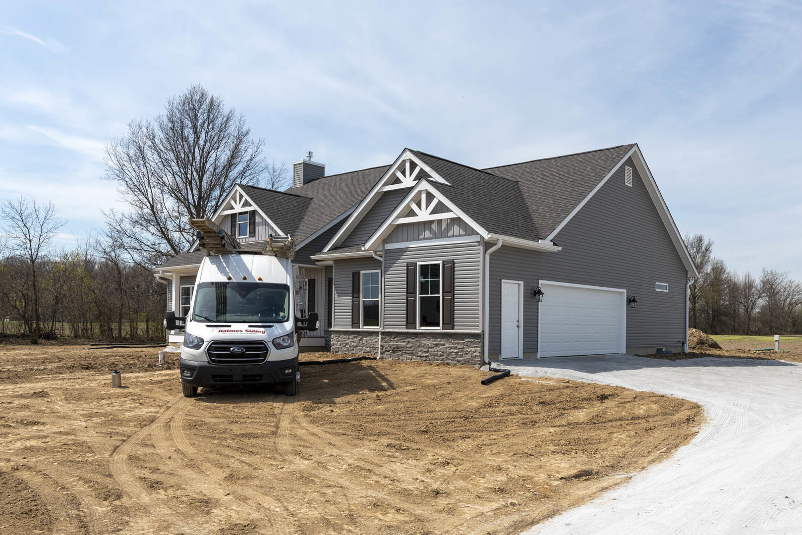 White pickup truck with ladder parked on driveway in front of house with white garage door, lamp post, and window framed in white trim; gray shingle roof and cloudy sky visible