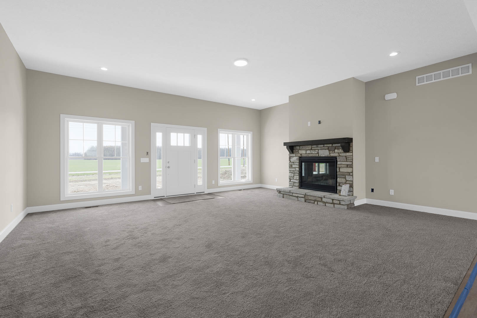 Living room with beige carpet, white walls, glass-front fireplace, large window overlooking a grassy field, and white door with glass panels