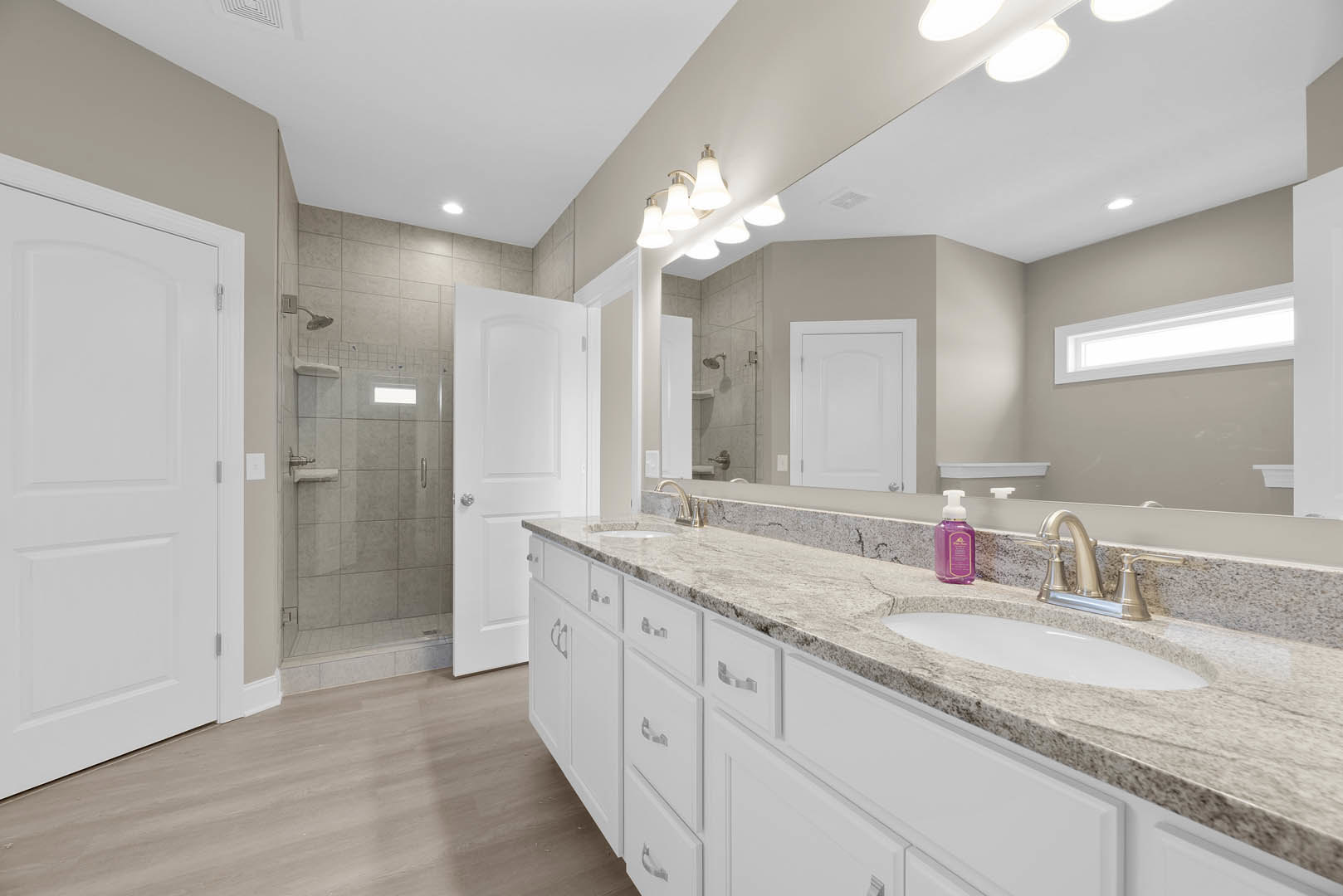 Bathroom with double white sinks, wide frameless mirror, wood flooring, white cabinetry, black and silver door handles, purple soap bottle on countertop