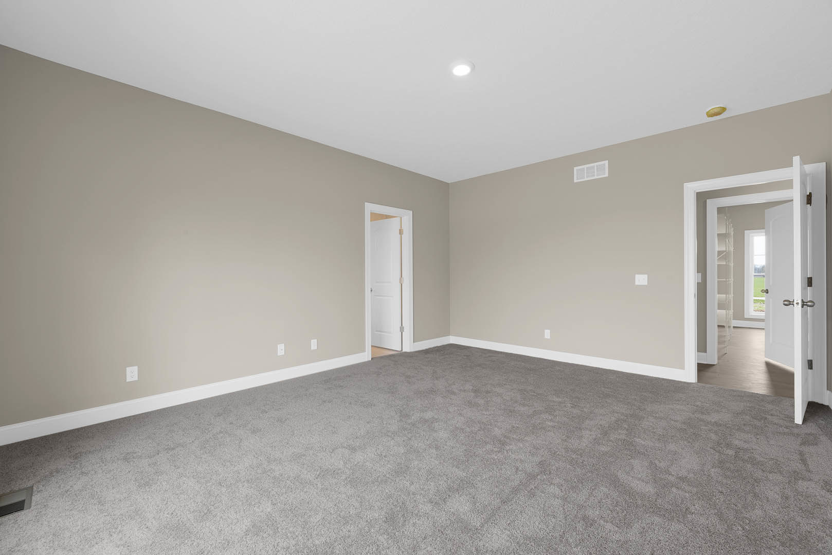 Grey carpeted room with white walls, open white door featuring wooden handle, white vent visible on wall, smooth plaster ceiling.