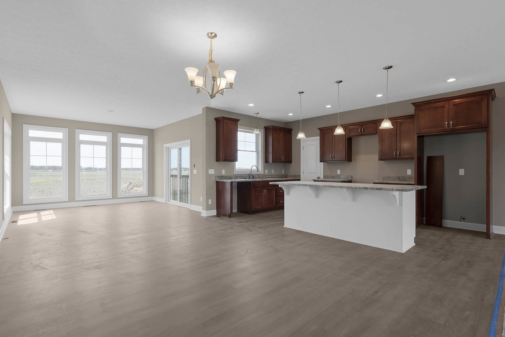 Open kitchen and living room featuring wood cabinets, wood flooring, a large countertop, row of windows overlooking a field, and a chandelier with white shades.