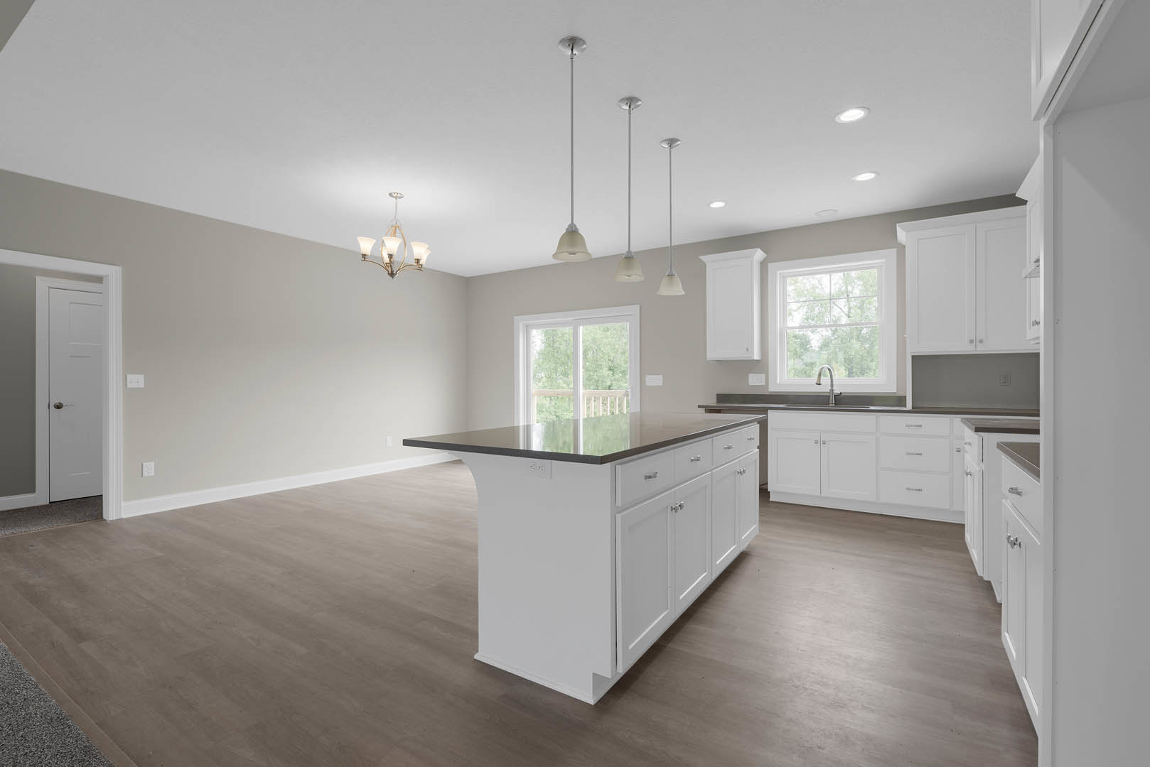 Kitchen with wood flooring, white cabinetry island featuring black countertop, stainless steel sink under window overlooking trees, metal support pole adjacent to island