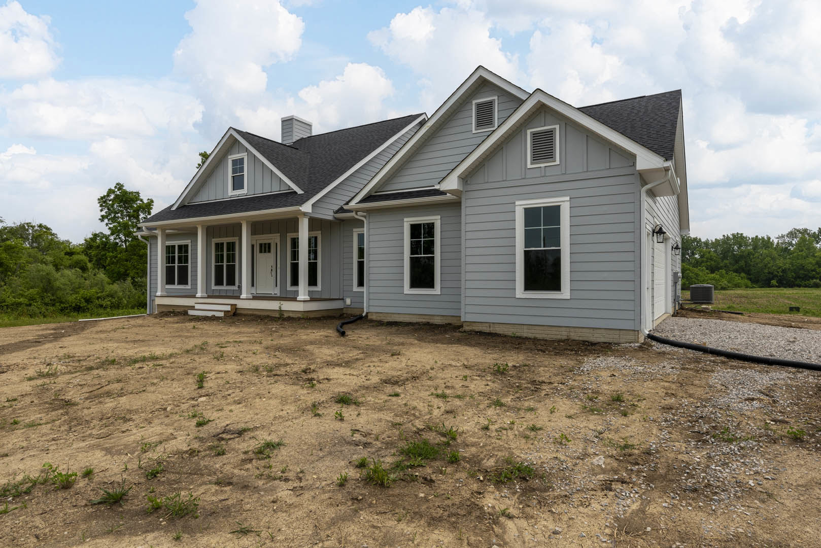 Grey house with white trim, white-framed windows, wall vents, and a dirt yard in front; cloudy sky and trees in background.