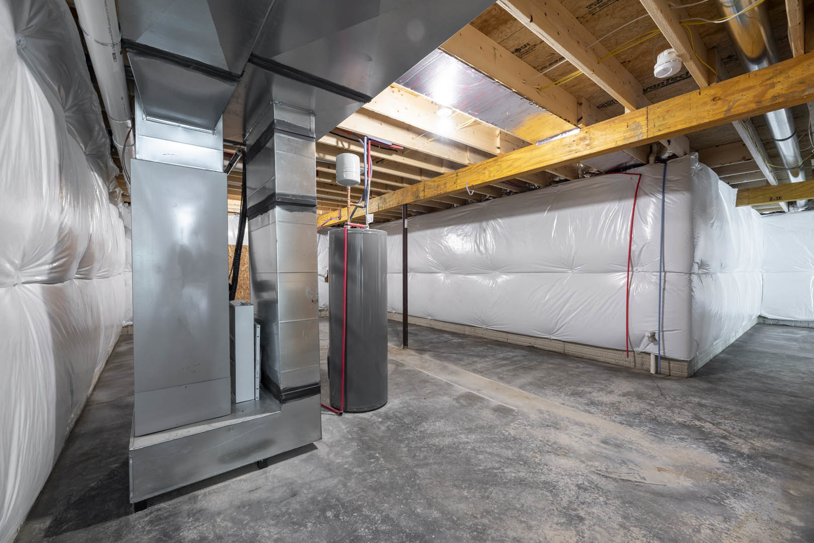 Basement room with expansive white wall, exposed concrete floor, metal tanks and containers, white cylinder on wood surface, smoke detector, large white bag mounted on wall, steel