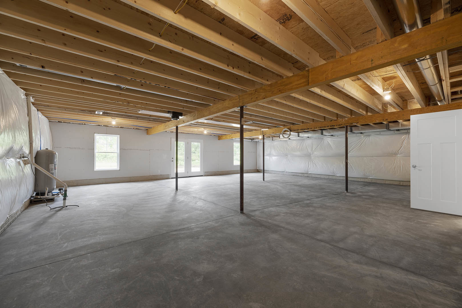 Concrete floor with exposed wooden ceiling beams, white-framed window, white door with silver handle, vertical support poles, and walls covered in white plaster or insulation