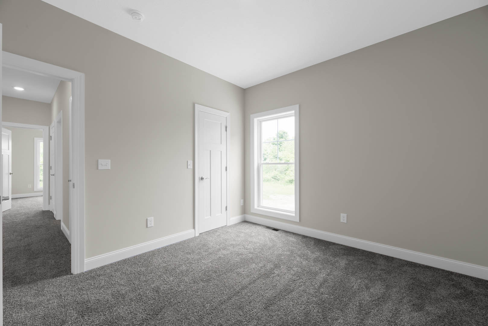 White-walled room with grey carpet, white door featuring a silver handle, and window overlooking green trees
