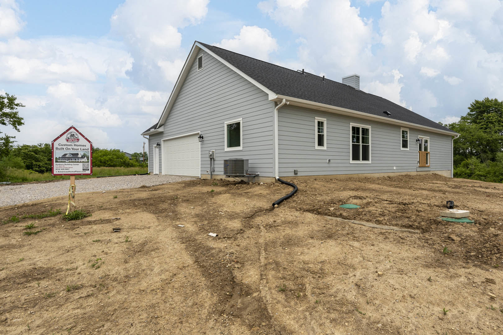 Grey house with black roof, large front window, sign on post in yard, black pipe in dirt, metal container on wheels, surrounded by trees and cloudy sky.