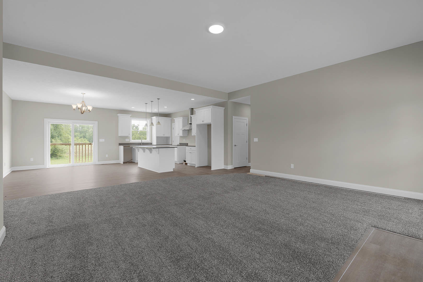 Open-concept living area with grey carpet, adjacent kitchen featuring white cabinetry and tile flooring, ceiling light fixture, window with wooden railing, and white door with
