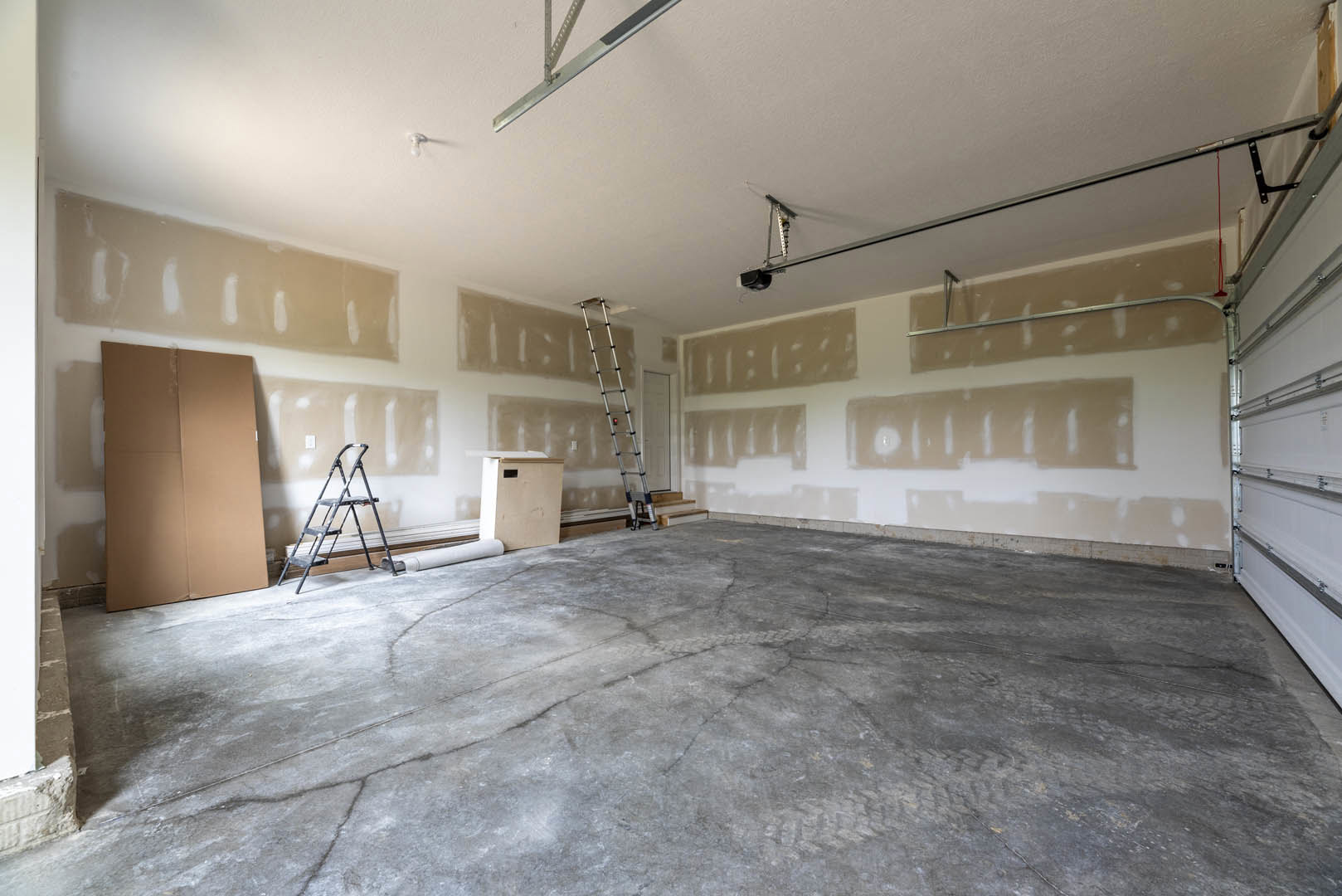Unfinished room with plaster walls, concrete floor showing tire tracks, metal ladders including one with a handle and red light, cardboard box, and white box with black label.