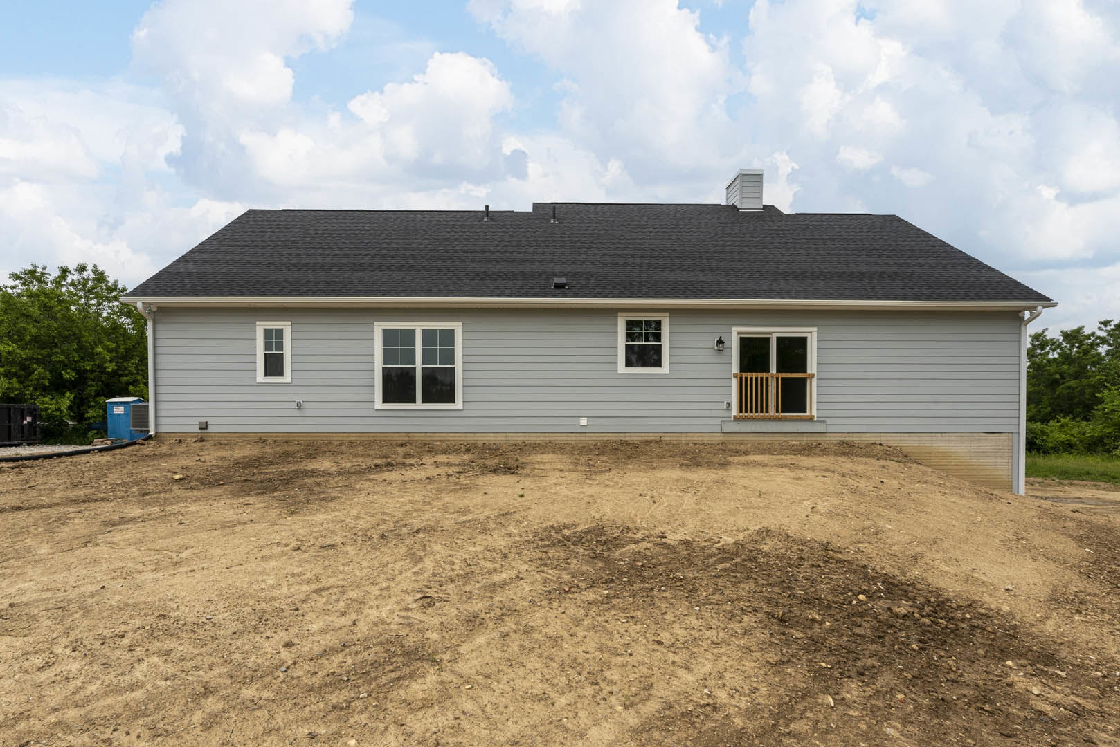 Modern house with white-framed windows, gray exterior walls, and a dirt lot in front, large green-leafed tree on the side, cloudy sky overhead