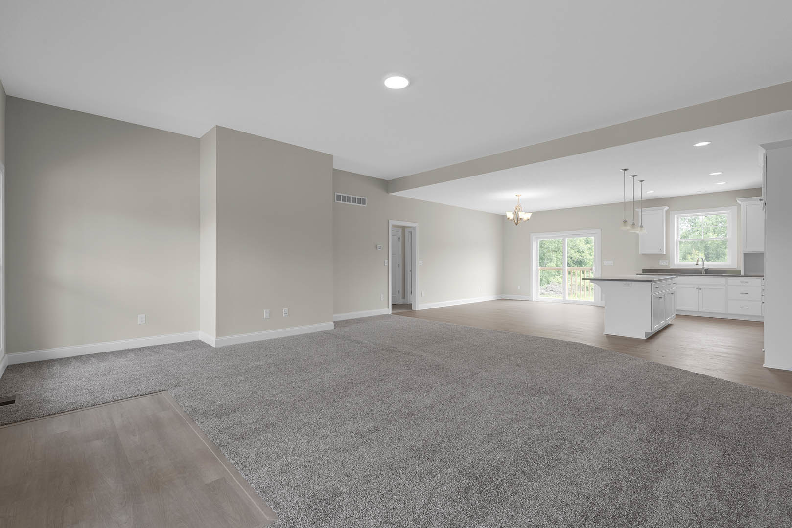 Spacious open-plan room featuring a modern kitchen with white cabinetry, large window overlooking trees, grey wood flooring with white trim, and a white door.