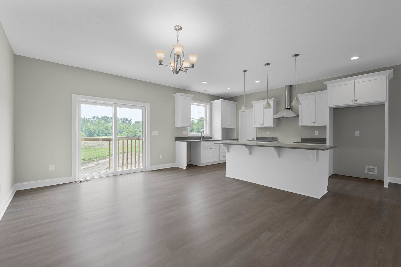 Open kitchen and dining area featuring hardwood floors, white cabinetry with grey countertops, sliding glass door overlooking a field and trees, and a modern chandelier above the
