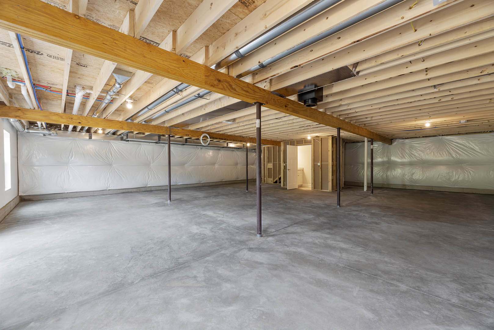 Exposed wooden ceiling beams, concrete floor with a metal support pole, visible pipes, white plastic sheeting covering part of the wall in an unfinished basement room