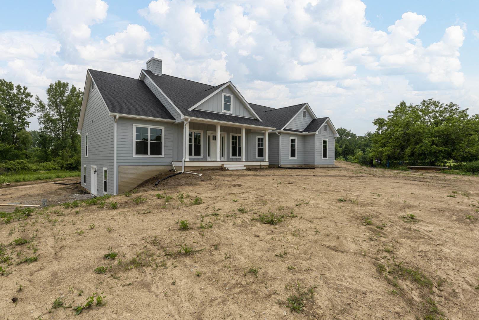 Two-story house with white-framed windows, gray roof, and large dirt hill in the yard, surrounded by young plants and trees under a cloudy sky