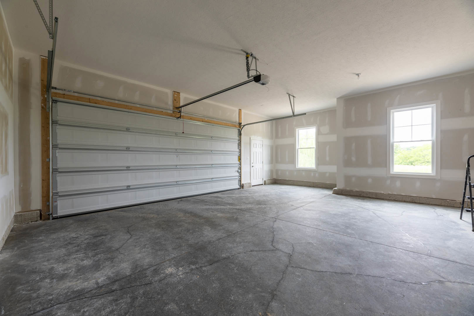 Concrete-floored room with a cracked surface, garage door framed in wood, white-framed window overlooking trees, and a white door with a doorknob; plaster walls and ceiling.