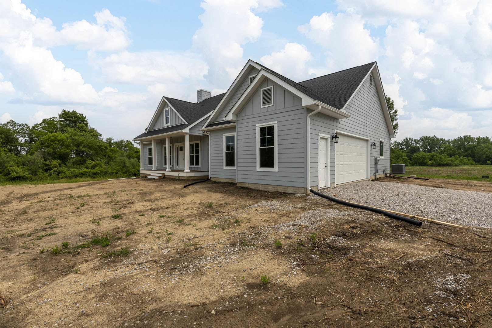 Grey cottage-style home with white-framed windows, white door, and multiple chimneys; driveway bordered by dirt patch with garden hose; mature trees and Robert Frost Farm visible
