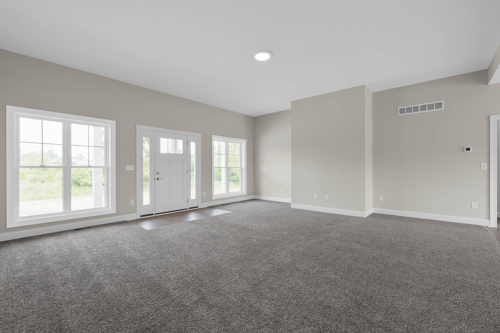 Carpeted room featuring large white-framed windows, white glass-paneled door, wall vent, and smooth plaster walls.