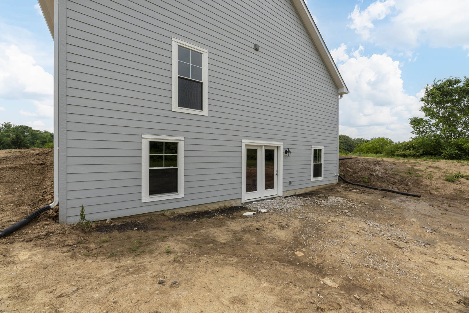 White double door with glass panels and adjacent windows set in light-colored siding, dirt patch and leafy tree in foreground, cloudy sky above