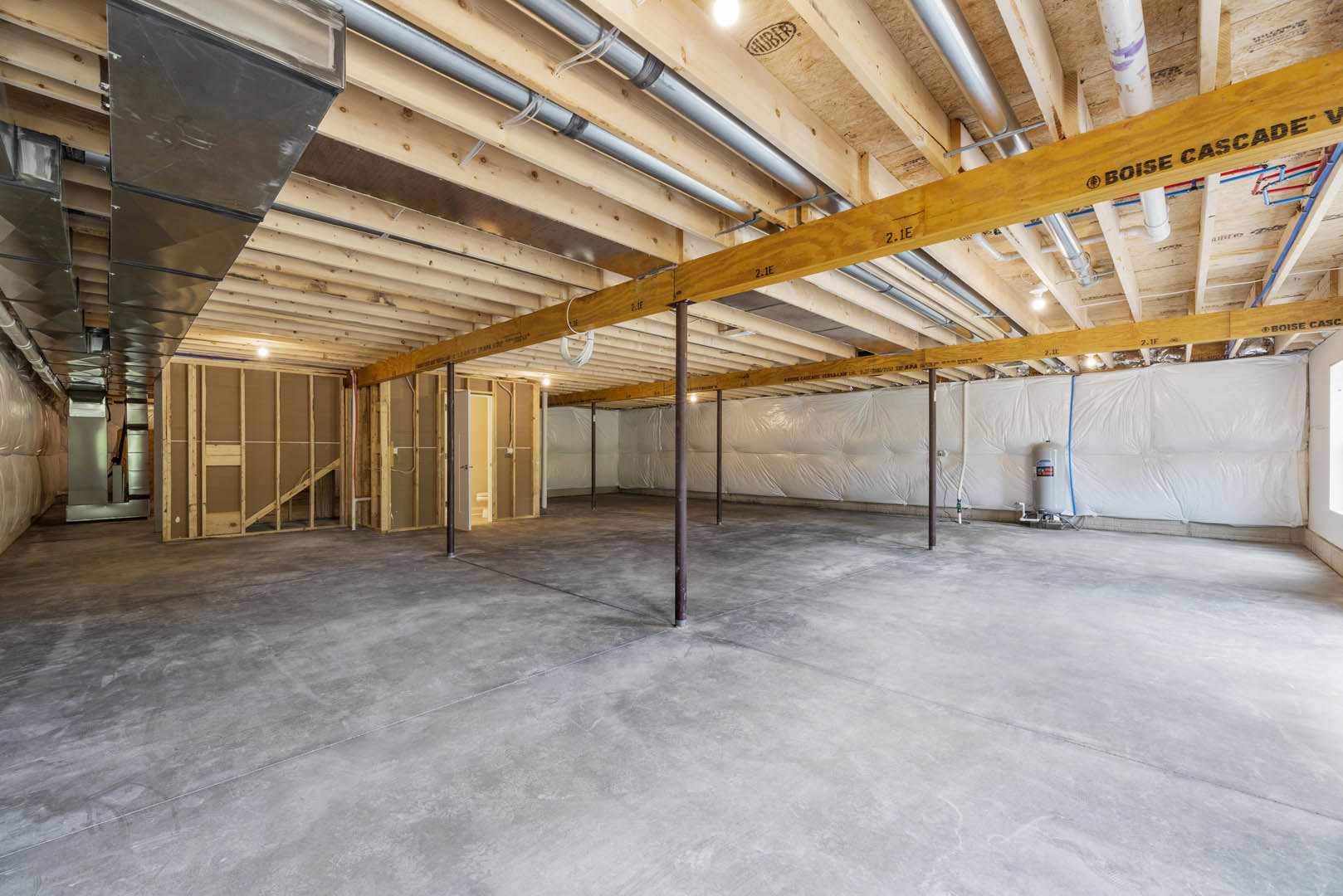 Exposed wood ceiling beams, concrete floor, white sheet covering one wall, visible pipes and structural supports in unfinished basement interior