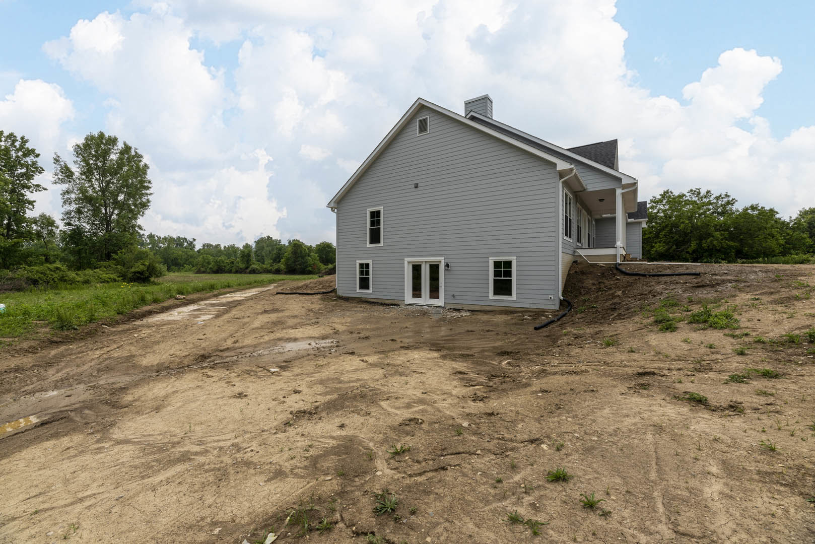 Grey house with white-framed windows and chimney, set on a steep dirt hill surrounded by tall leafy trees; person standing near the entrance.