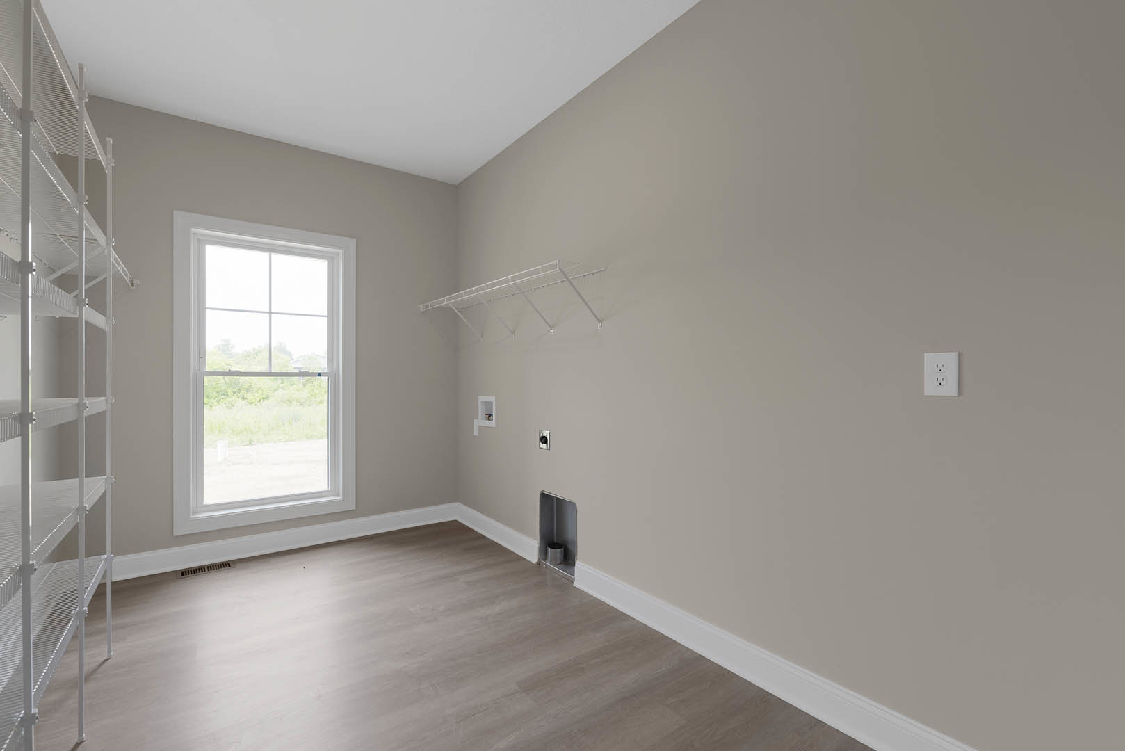 Bright room with laminate wood flooring, white plaster walls, large window overlooking outdoors, white metal shelf with railing, silver trash can in corner, and white electrical