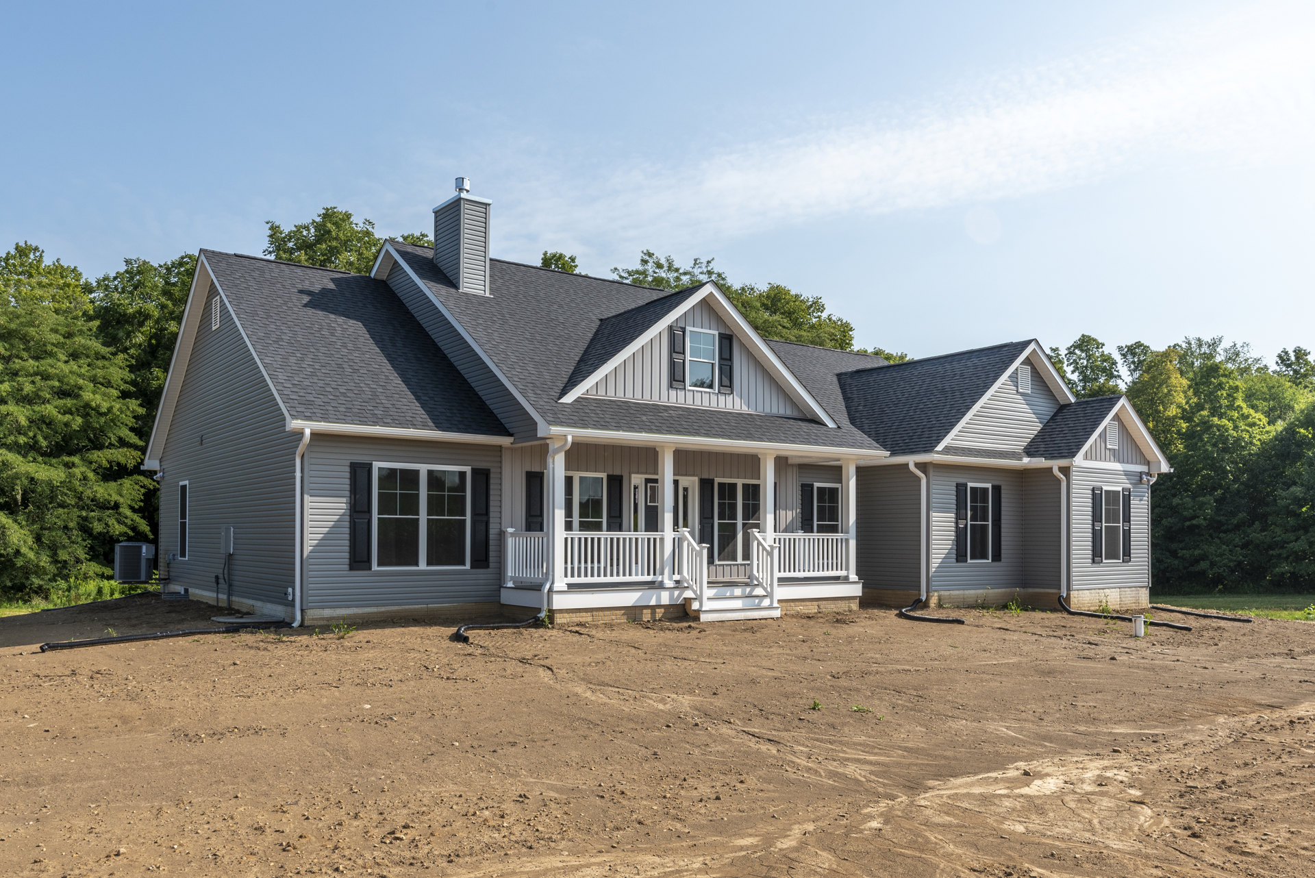 Two-story house with gray siding, white-trimmed windows, and a porch with white railing, set on a dirt yard; mature trees and Robert Frost Farm visible in the background.