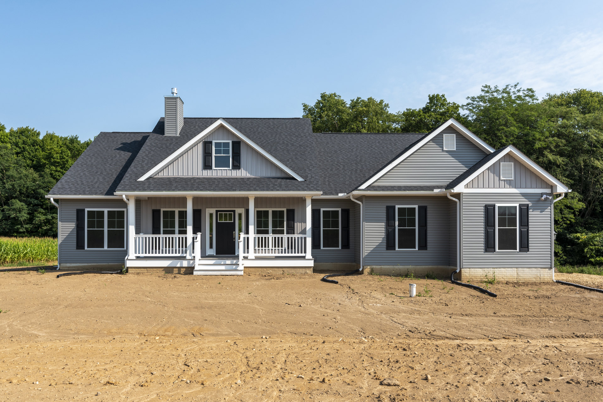 Two-story house with black shuttered windows and door, white porch railing, dirt yard featuring a black pipe emerging from the ground