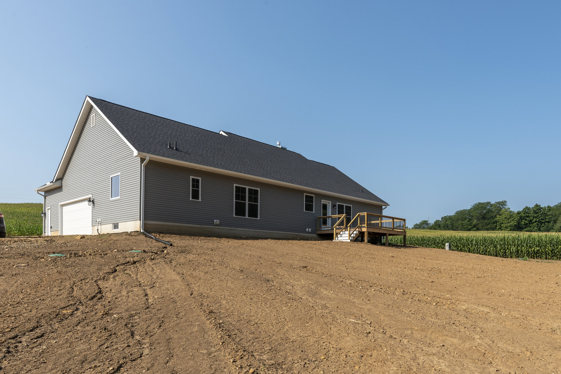 Two-story house with gray siding and gabled roof, wooden deck with railings, large dirt field and sparse trees in rural setting