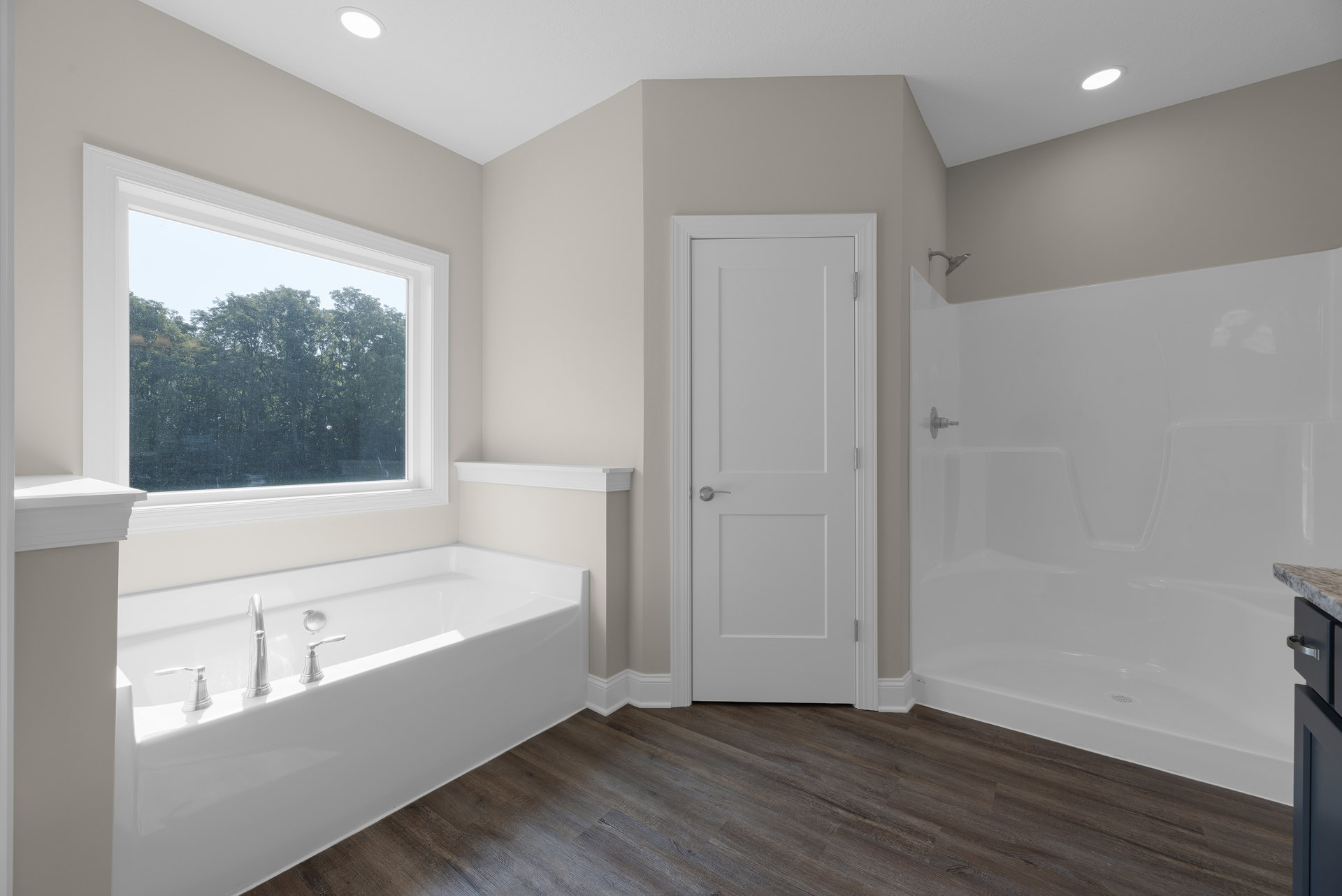 Bathtub with chrome faucets set on wood flooring, white door with silver handle, large window overlooking trees, white shower enclosure, plaster walls, cabinetry.