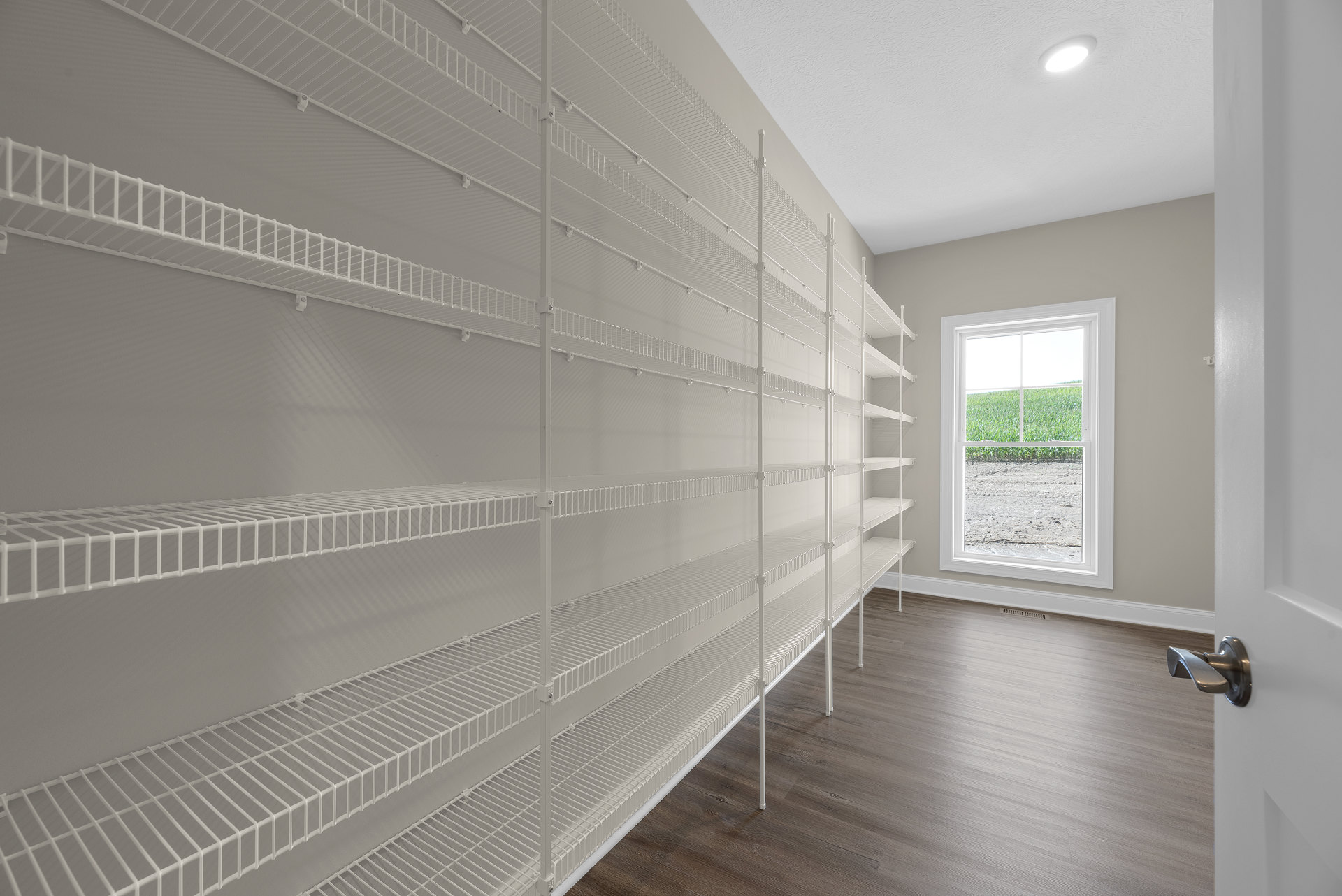 Built-in white shelves along a plaster wall, large window overlooking grassy field, marble tile flooring, recessed ceiling light, close-up of brushed metal door handle