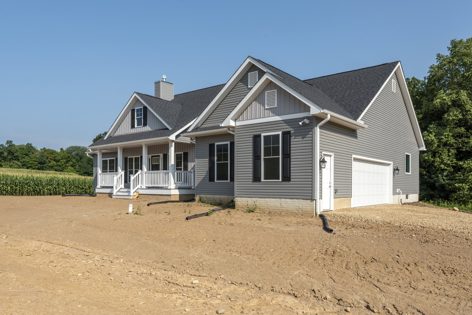 Two-story house with white siding, black shuttered windows, white framed windows, and white railing; dirt field in foreground with black pipe on ground; trees and blue sky in