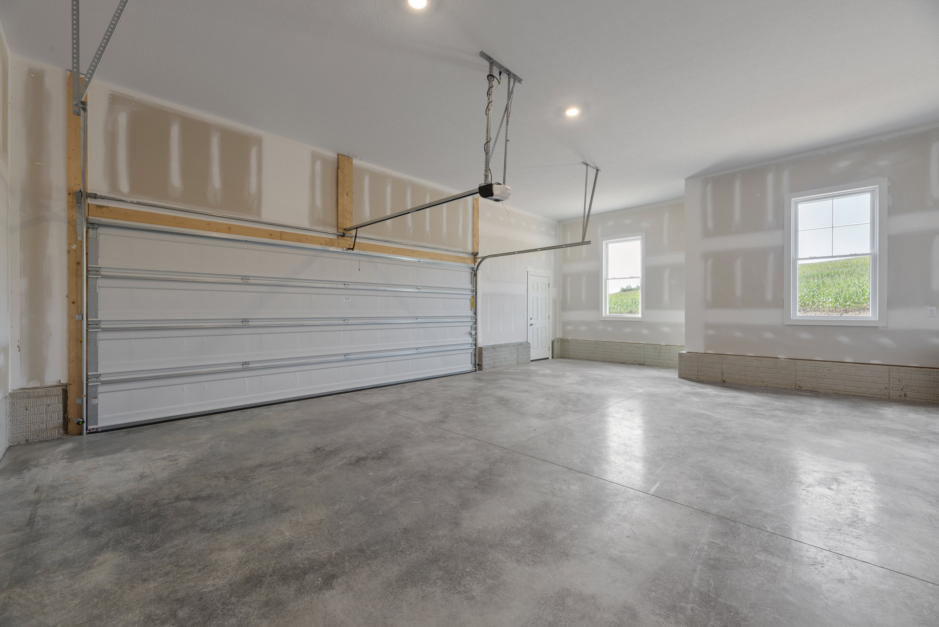 Garage interior with smooth concrete floor, white paneled garage door framed by a wooden beam, plaster walls, ceiling, and a window overlooking a grassy field