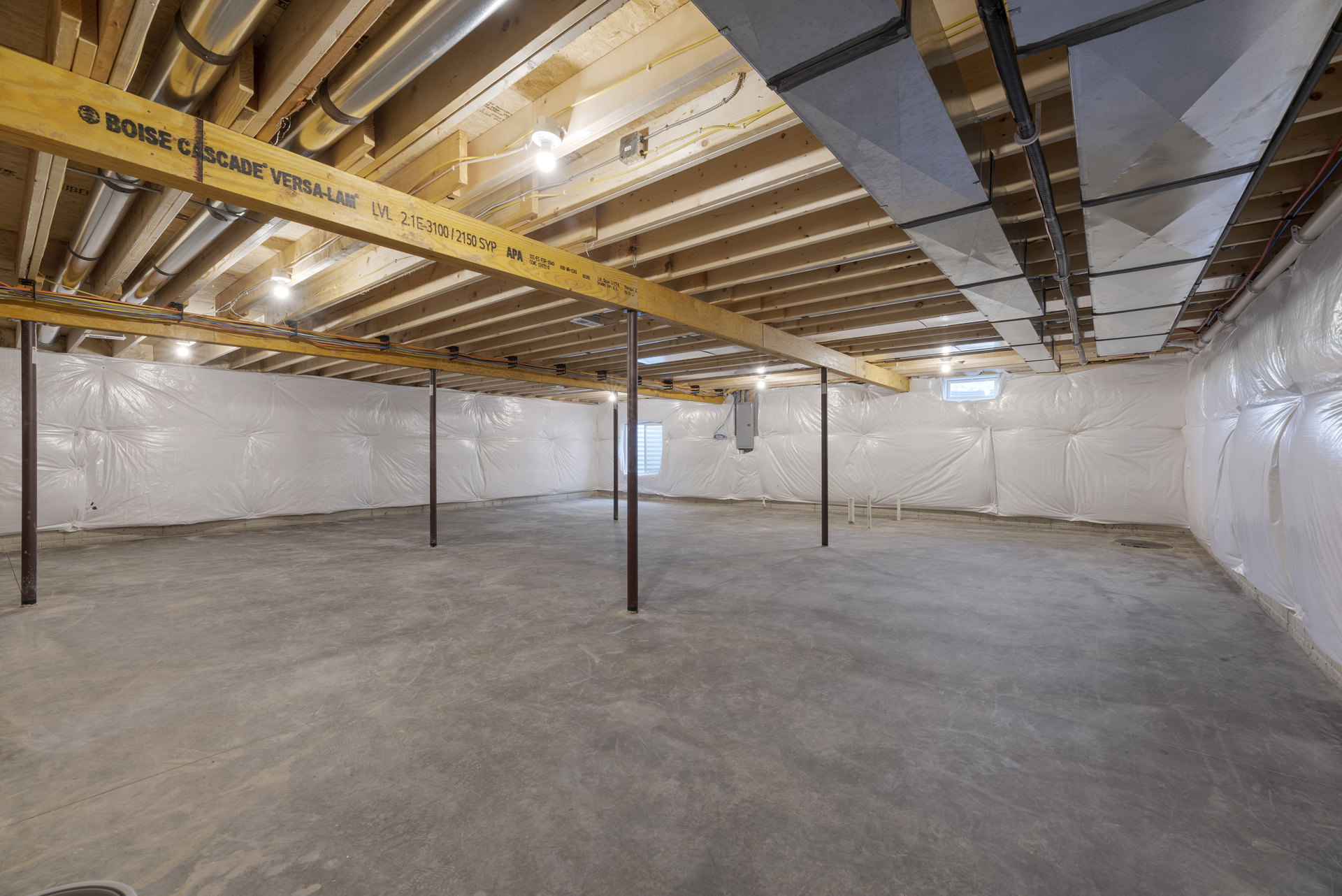 Basement room with exposed wooden and metal beams, white walls and ceiling, concrete floor, ceiling-mounted metal pipes, and a hanging light fixture.