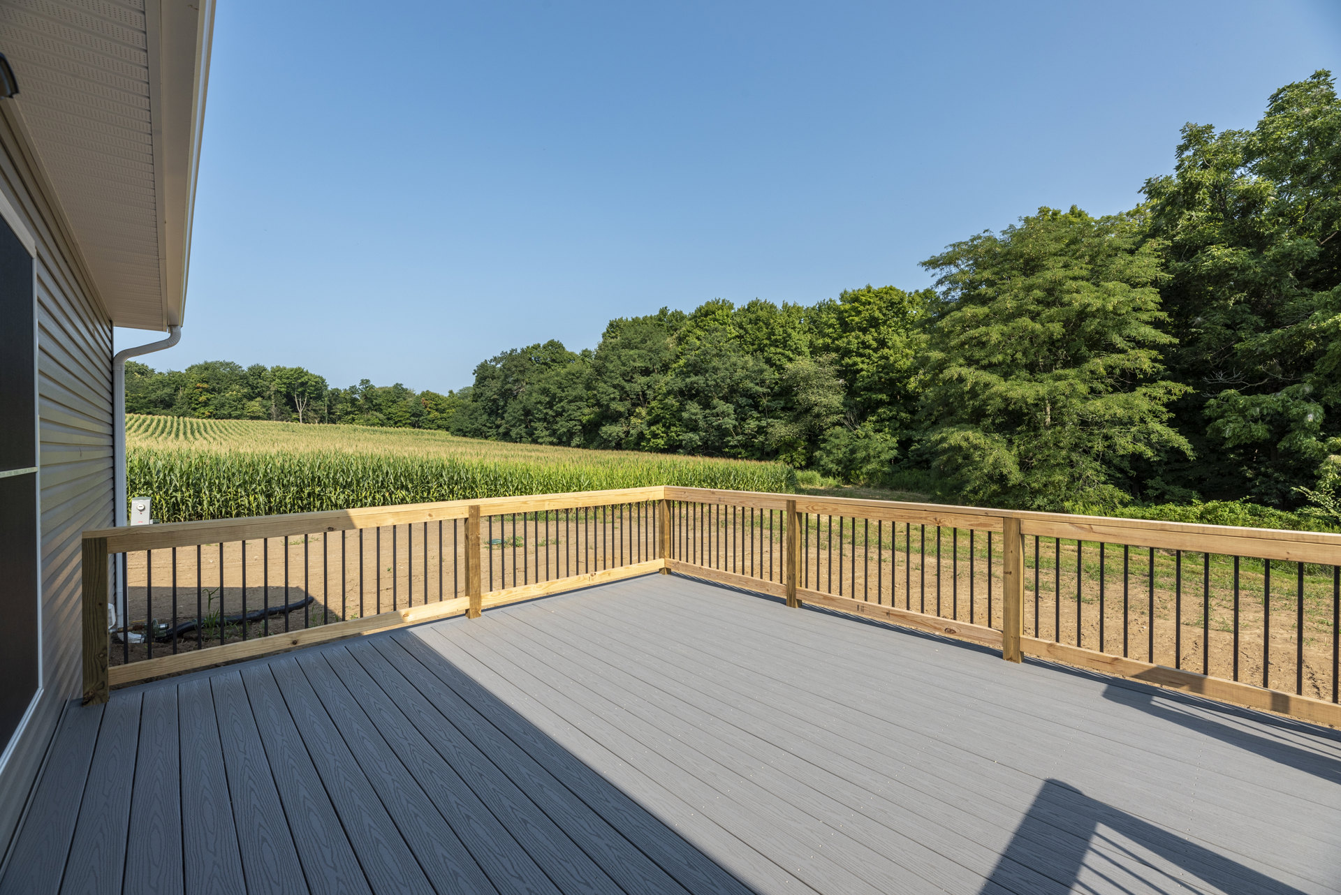 Wooden deck with railing, bordered by a fence, overlooking grassy yard and mature trees under blue sky