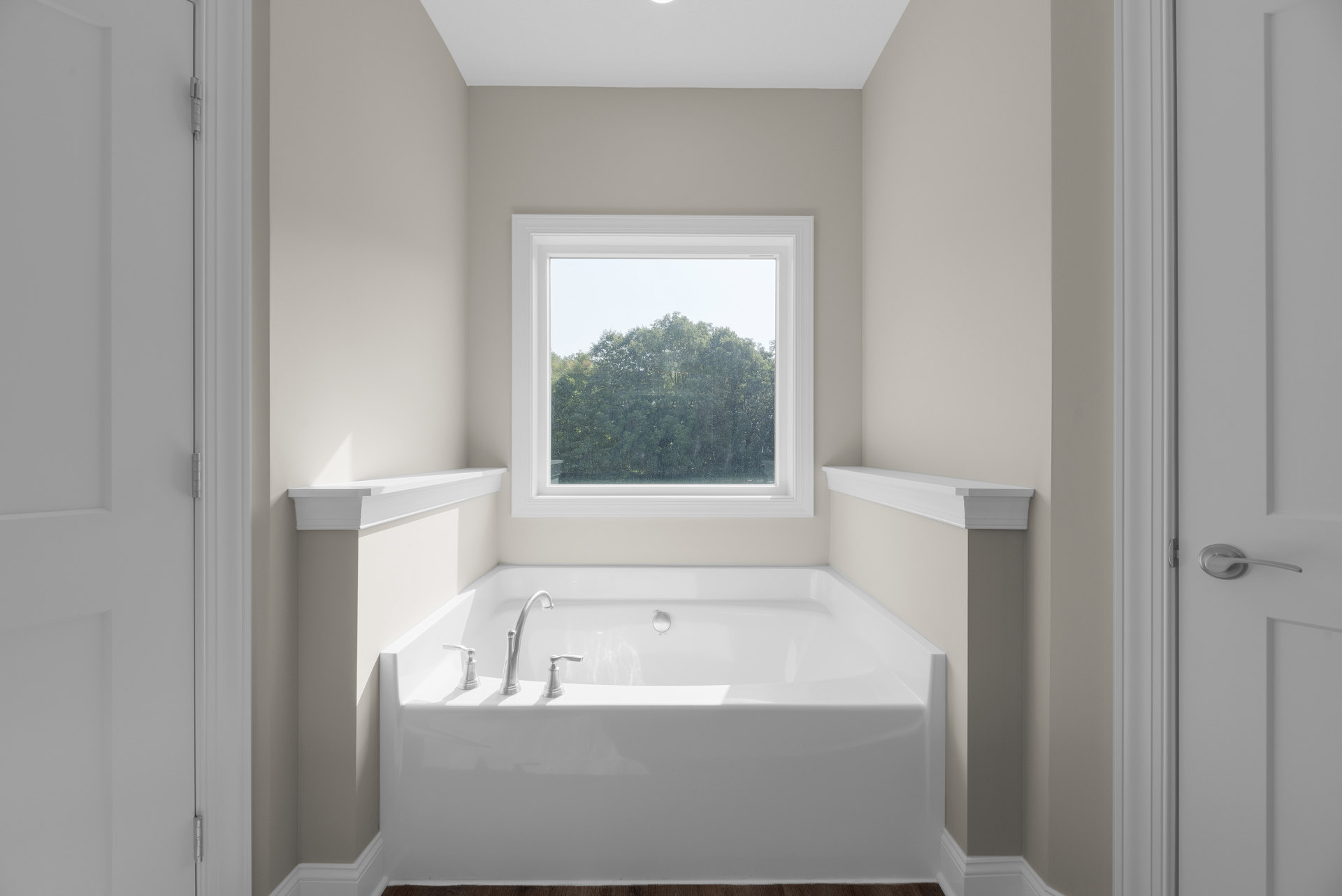 Freestanding white bathtub with chrome faucet beneath large window overlooking leafy trees, white shelf mounted on wall, minimalist bathroom design with black and white finishes