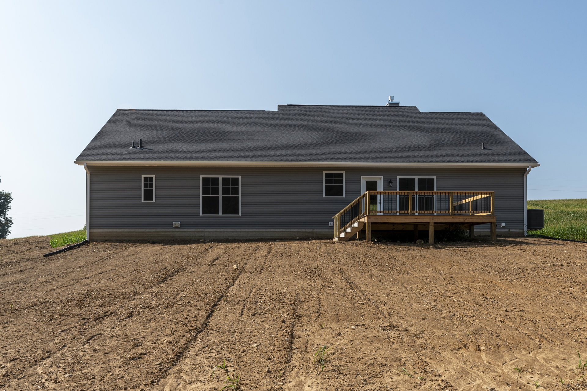 Wooden deck with white railing attached to beige house, large window with white trim, dirt field with tire tracks in foreground, sparse vegetation.
