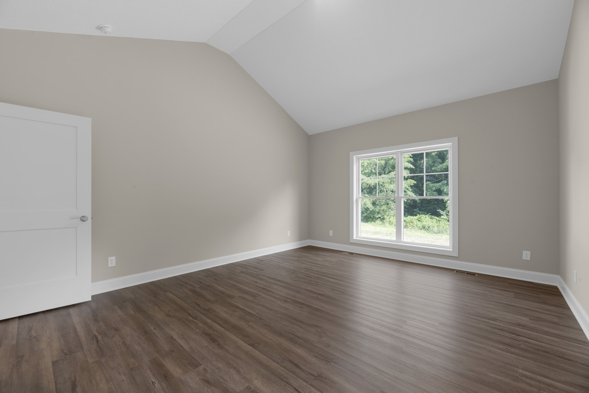 Sunlit room featuring a large window overlooking green trees, hardwood flooring, white walls, and a white door with a silver handle