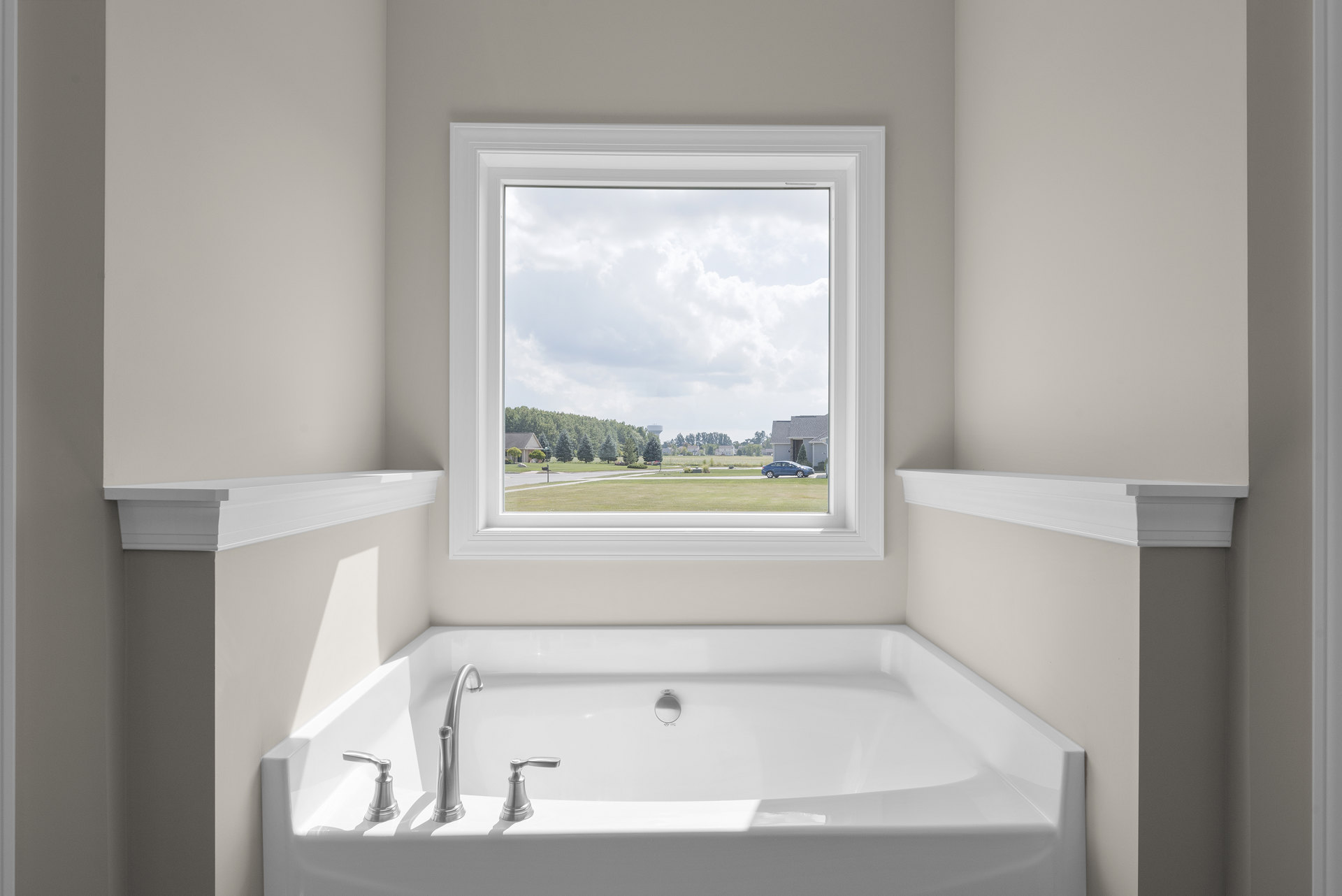 Freestanding white bathtub with silver faucet beneath large window, white wall molding, parked car visible outside