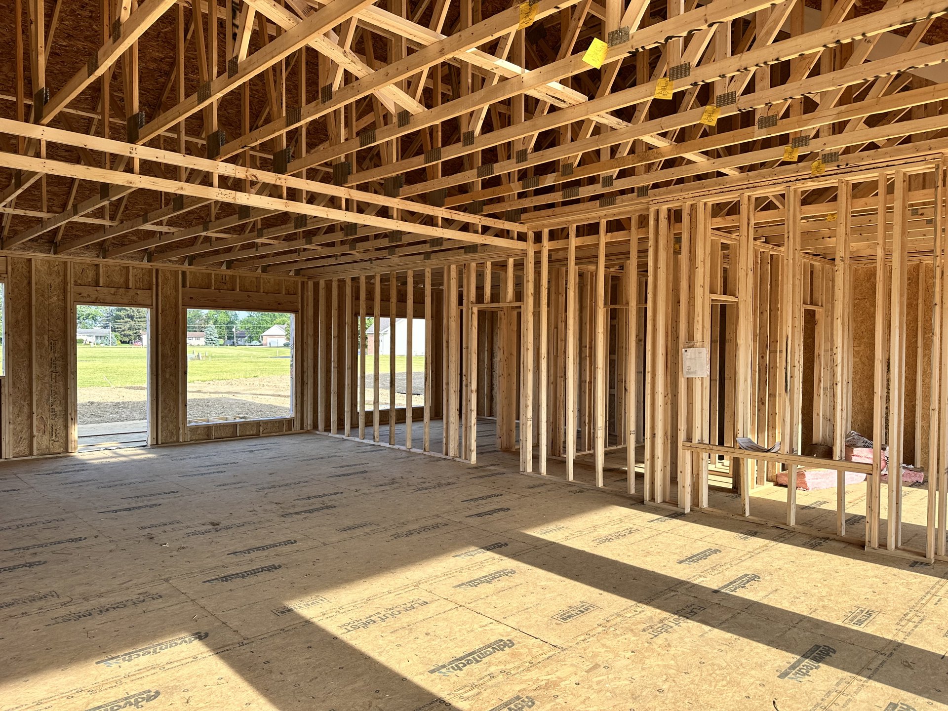 Wooden house frame under construction with exposed beams, plywood flooring, yellow tape on roof structure, and window overlooking a field and trees