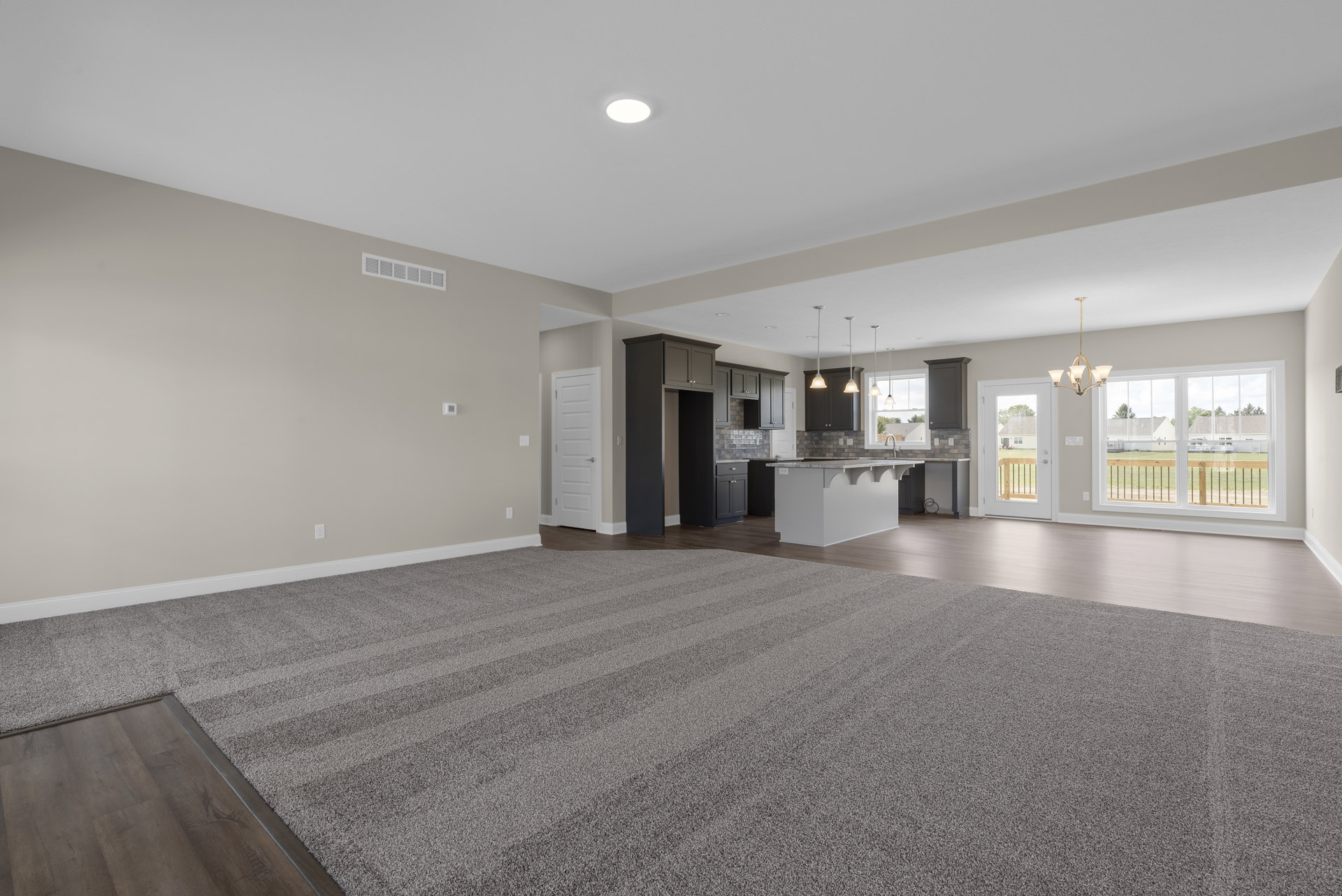 Spacious living room with plush carpet, open kitchen featuring white cabinetry and stone island, dining area, white door with silver handle, modern chandelier, mix of laminate and