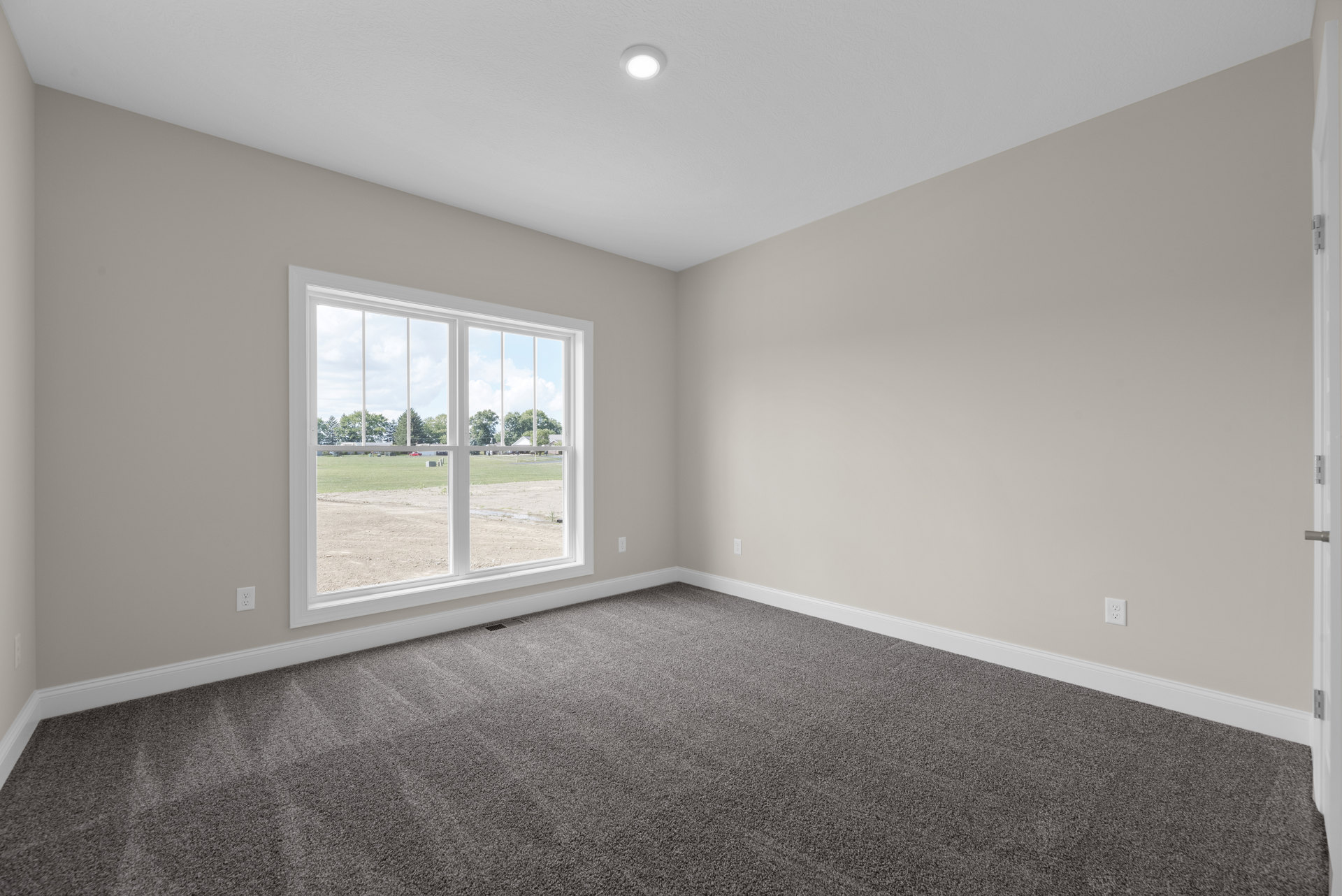 Bright room with white walls, large window overlooking trees and field, grey carpet flooring, recessed ceiling light, simple molding.