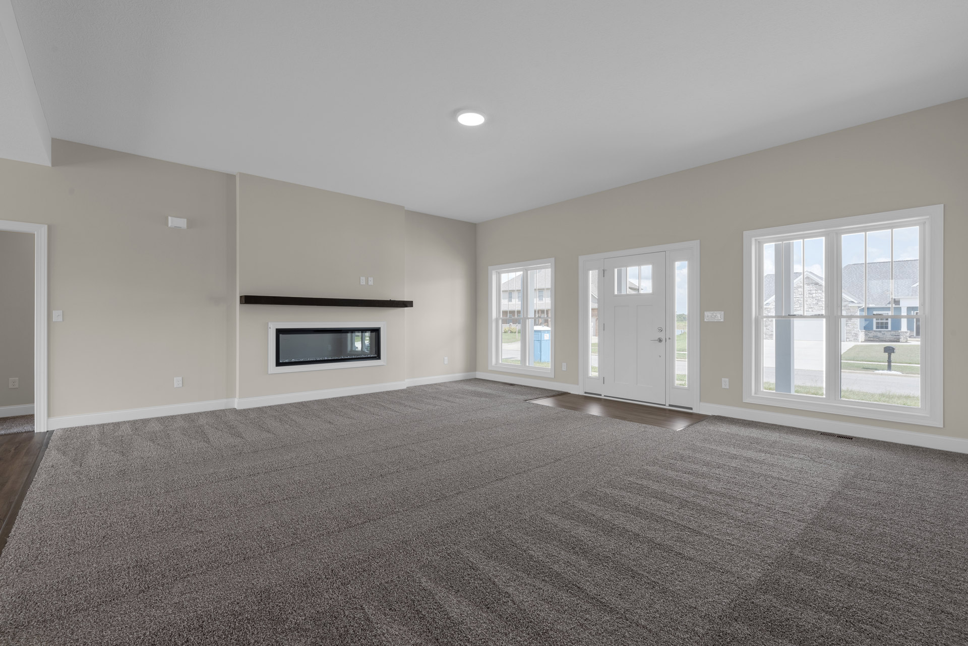 Living room with beige carpet, white plaster walls, glass-front fireplace, large windows showing lawn and neighboring house, and a white door with glass panes.