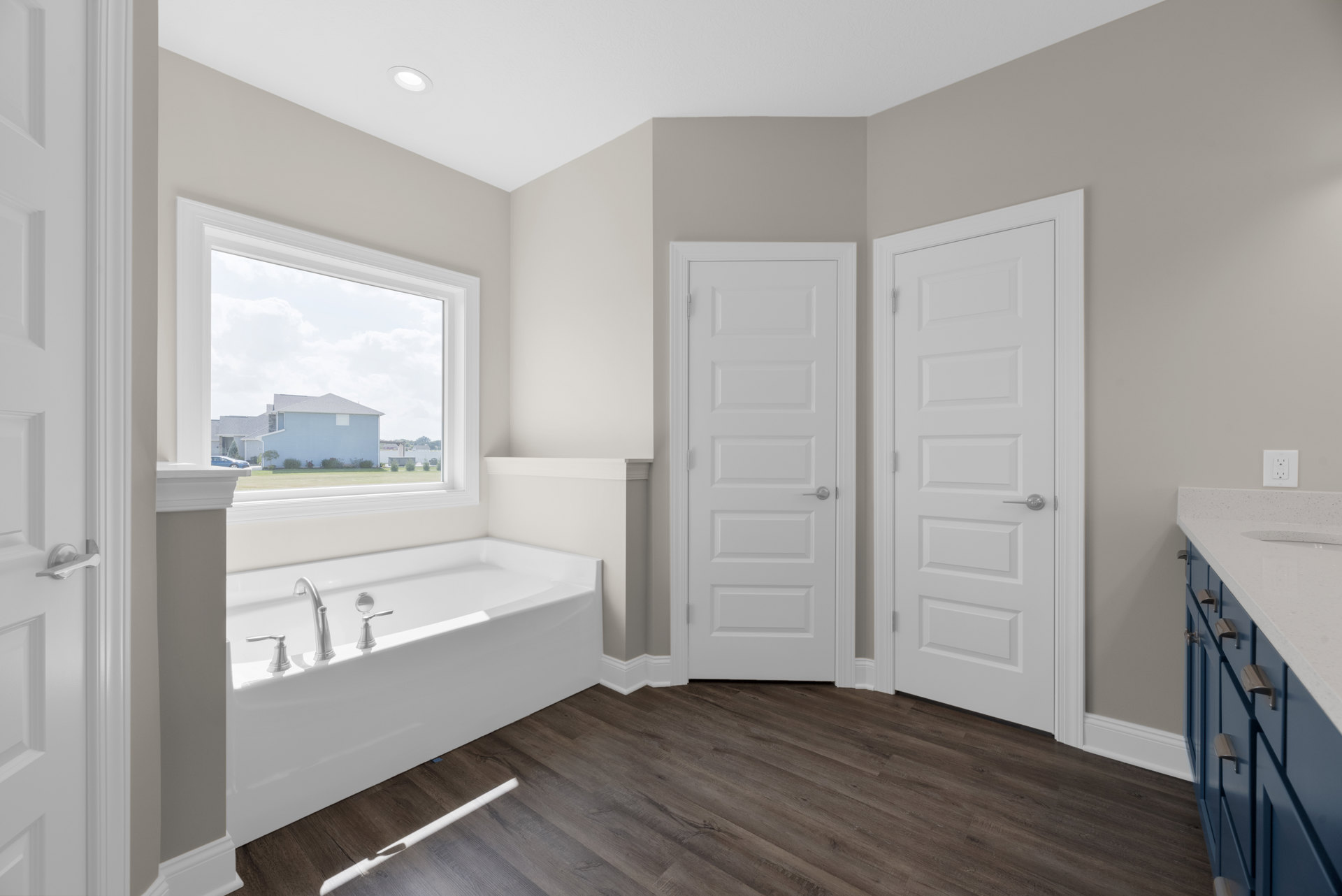 Bathroom with freestanding bathtub, silver faucets, wood flooring, white trim, white door with silver handle, window overlooking blue house and green lawn